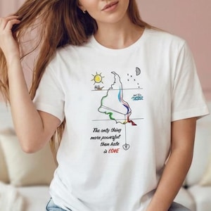 May include: White t-shirt with a colorful graphic and the text "The only thing more powerful than hate is LOVE." The graphic includes a sun, moon, and a stylized shape with rainbow colors. The model is wearing blue jeans.