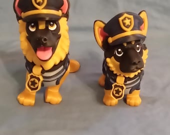 May include: Two toy German Shepherd dogs wearing police hats and badges. The dogs have black and yellow fur, black noses, and pink ears. They are posed on a light blue surface.