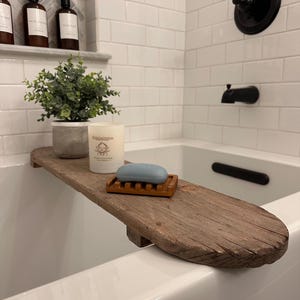 May include: A rustic wooden bathtub tray holds a potted plant, a scented candle, a wooden soap dish, and a blue bar of soap. The tray rests on the edge of a white bathtub. The background features white subway tiles and black fixtures.