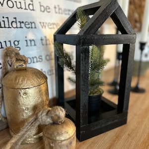 May include: A black wooden lantern with a small potted evergreen tree inside. Beside the lantern are two gold-colored bells with rope hangers. A white sign with black text is in the background.