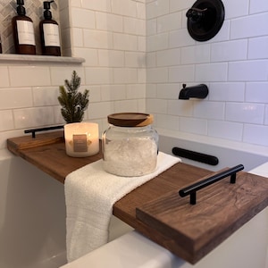 May include: A dark wood bathtub tray with black handles, holding a white towel, a lit candle, and a glass jar with a wooden lid. The tray sits across a white bathtub, with a black faucet and shower fixtures.