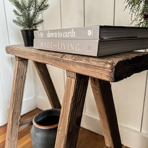 May include: A rustic, wooden stool with a distressed finish. Two books, "down to earth" and "MADE for LIVING", are stacked on top. A small potted evergreen and a dark ceramic pot are also visible.