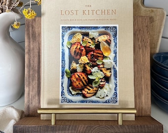 May include: A cookbook titled "The Lost Kitchen" with a photo of grilled food on a blue and white plate. The book is displayed on a dark wood shelf with a brass handle. Yellow flowers in a white vase are in the background.