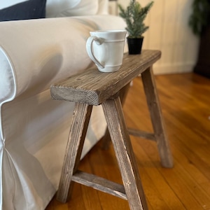 May include: A small, rustic wooden side table with a distressed finish. A white ceramic mug with a curved handle sits on top, next to a small potted evergreen. The table has a simple, A-frame leg design, perfect for a living room or bedroom.