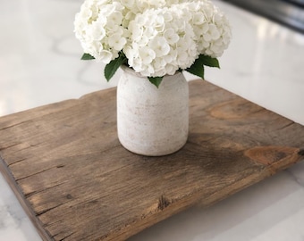 May include: A rustic wooden serving tray with a distressed finish holds a small, off-white vase filled with white hydrangea flowers. The tray sits on a white marble countertop, creating a contrast of textures and colors. The arrangement suggests a farmhouse or cottagecore aesthetic.