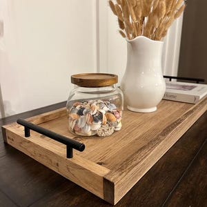 May include: A wooden serving tray with black handles holds a glass jar filled with seashells and a white vase with dried flowers. The tray is made of light brown wood.