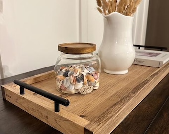 May include: A wooden serving tray with black handles holds a glass jar filled with seashells and a white vase with dried flowers. The tray is made of light brown wood.