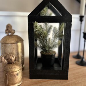 May include: A black wooden lantern with a small potted evergreen tree inside. Two antique gold-colored bells and a larger gold-colored container are to the left of the lantern. The objects are on a wooden surface.