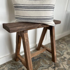 May include: A rustic, wooden bench with a rectangular seat and angled legs. A cream-colored pillow with horizontal navy blue stripes rests on the bench. The bench is made of dark brown wood and sits on a patterned rug.