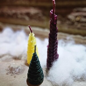 May include: Three decorative candles in a variety of colors. A dark green candle is in the foreground, a yellow candle is in the middle, and a tall, textured, deep purple candle is in the background. The candles are on a light brown surface with white fluff.