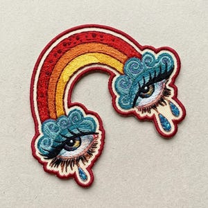 May include: An embroidered patch featuring a curved rainbow in shades of red, orange, and yellow. At each end of the rainbow, a stylized eye is nestled within blue cloud-like shapes, with long black eyelashes and a single blue teardrop falling.