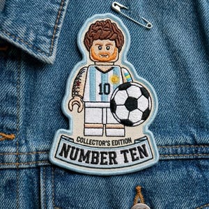 May include: An embroidered patch featuring a stylized figure resembling a toy brick character with brown hair and a beard, wearing a light blue and white striped soccer jersey with the number 10, holding a soccer ball, with text reading COLLECTOR'S EDITION NUMBER TEN.