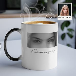 May include: White ceramic mug with a black handle, featuring a black and white image of eyes and the text "Can't take my eyes off you." The mug is filled with a hot beverage, and steam rises from the top. A small photo is in the upper right corner.
