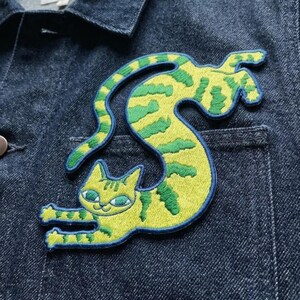 May include: An embroidered patch featuring a yellow cat with green stripes, depicted in a long, curved, stretching pose. The cat has green eyes and is stitched with a dark blue border, attached to the front pocket area of a dark blue denim jacket.