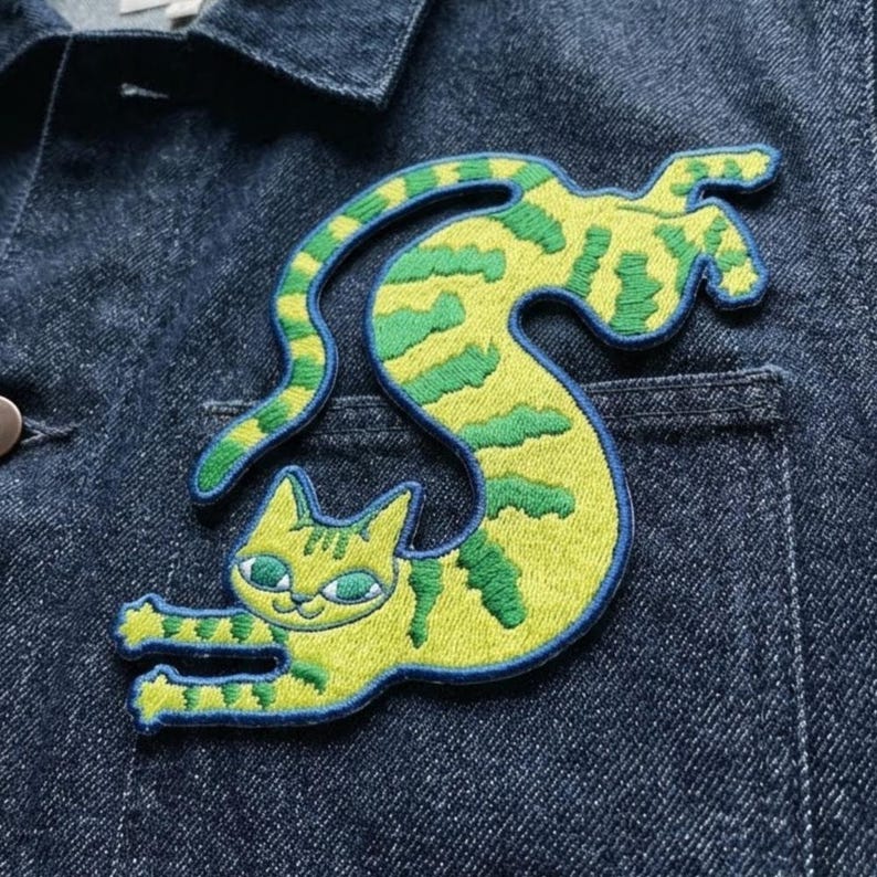 May include: An embroidered patch featuring a yellow cat with green stripes, depicted in a long, curved, stretching pose. The cat has green eyes and is stitched with a dark blue border, attached to the front pocket area of a dark blue denim jacket.