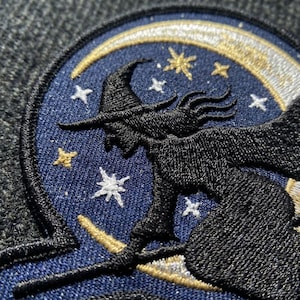 May include: A circular embroidered patch featuring a black silhouette of a figure wearing a pointed hat and riding a broomstick against a dark blue background. The design includes a gold crescent moon and several white and gold embroidered stars.