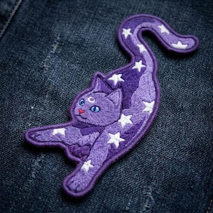 May include: An embroidered patch shaped like a stretching cat in shades of purple. The cat features a small white crescent moon on its forehead, bright blue eyes, a red nose, and is decorated with numerous white stars across its body and tail.