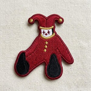May include: An embroidered patch featuring a jester figure in a deep red suit with a matching two-pointed hat. The figure has a white face with red markings, a gold collar, three gold buttons down the front, and solid black feet.