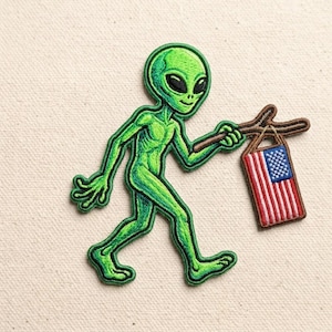 May include: An embroidered patch featuring a bright green alien with large black eyes walking forward. The alien holds a brown stick over its shoulder, from which hangs a small rectangular flag with red and white stripes and a blue field with stars.