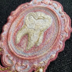 May include: An oval embroidered patch featuring a central off-white tooth motif surrounded by an ornate, scalloped frame in shades of pink, peach, and light blue. The edges of the frame are detailed with intricate stitching and gold-toned accents.