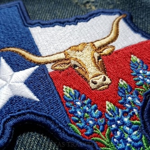 May include: A Texas-shaped embroidered patch featuring a blue section with a white star, a white section, and a red section. The design includes a tan longhorn steer head with long horns and a cluster of bluebonnet flowers with green stems.