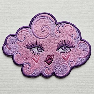 May include: An embroidered patch shaped like a cloud in shades of pink and purple. The cloud features a face with long eyelashes, dark purple eyes, a small puckered mouth, and two pink heart-shaped cheeks, all outlined in a deep purple border.