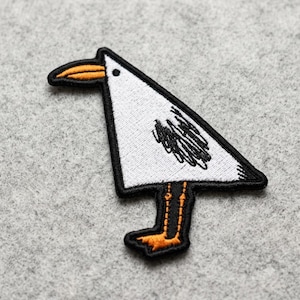 May include: An embroidered patch shaped like a stylized bird with a triangular white body, a black outline, and a black scribbled wing detail. The bird features an orange beak and orange legs with webbed feet, all rendered in a flat, graphic style.