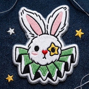 May include: An embroidered patch featuring a white rabbit head with long ears, pink inner ears, a red heart-shaped nose, and a yellow star over one eye. The rabbit wears a green and black ruffled collar, surrounded by small embroidered stars.