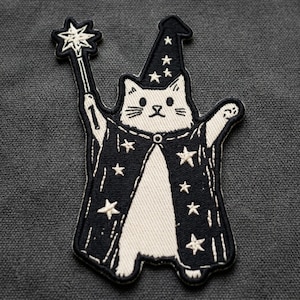 May include: An embroidered patch featuring a white cat dressed as a wizard. The cat wears a black pointed hat and a black cape, both decorated with white stars. The cat holds a white star-tipped wand in one raised paw, with a black border.