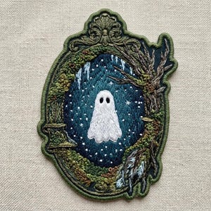 May include: An embroidered patch featuring a white ghost centered within a dark blue, starry night scene. The scene is framed by an ornate, olive green border decorated with mossy textures, bare tree branches, and intricate scrollwork at the top.