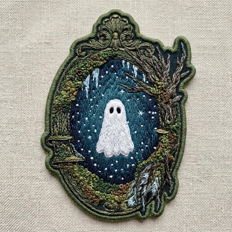 May include: An embroidered patch featuring a white ghost centered within a dark blue, starry night scene. The scene is framed by an ornate, olive green border decorated with mossy textures, bare tree branches, and intricate scrollwork at the top.