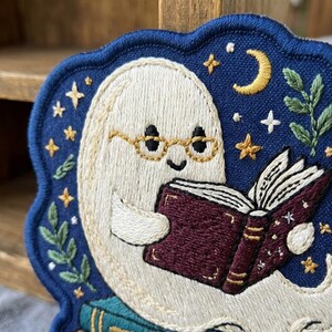 May include: An embroidered patch featuring a white ghost wearing gold-rimmed glasses and reading an open dark red book. The background is deep blue with yellow stars, a crescent moon, and green leafy sprigs. The patch has a scalloped dark blue border.