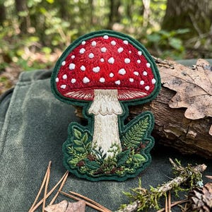 May include: An embroidered patch featuring a red mushroom cap with white spots, a cream-colored stalk, and green foliage at the base. The design is outlined in a dark green border, showcasing detailed thread work and a textured appearance.