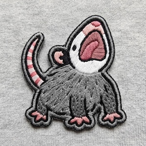 May include: An embroidered patch featuring a gray opossum with a white face, pink nose, and pink ears. The opossum has its mouth wide open, revealing a pink tongue, and a long, striped pink and white tail curled upward against a gray fabric.