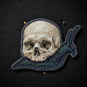 May include: An embroidered patch featuring a human skull with beige and white thread details, integrated into the body of a dark blue and green snail. The design is outlined in a dark gray border and secured with small gold-toned metal pins.