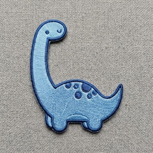 May include: A light blue embroidered dinosaur patch with a long neck and a dark blue border. The dinosaur features a series of dark blue circular spots along its back and a simple stitched eye and mouth detail on its rounded head.