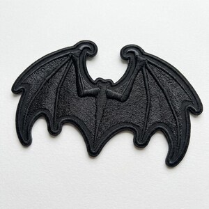 May include: A black embroidered patch in the shape of a bat with outstretched wings. The design features detailed stitching that defines the skeletal structure of the wings and the central body, with a scalloped edge along the bottom of the wings.