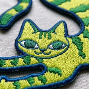 May include: An embroidered patch featuring a stylized cat with lime green fur and dark green stripes. The cat has large, almond-shaped green eyes with white accents and a dark blue stitched border that outlines the entire shape of the feline figure.