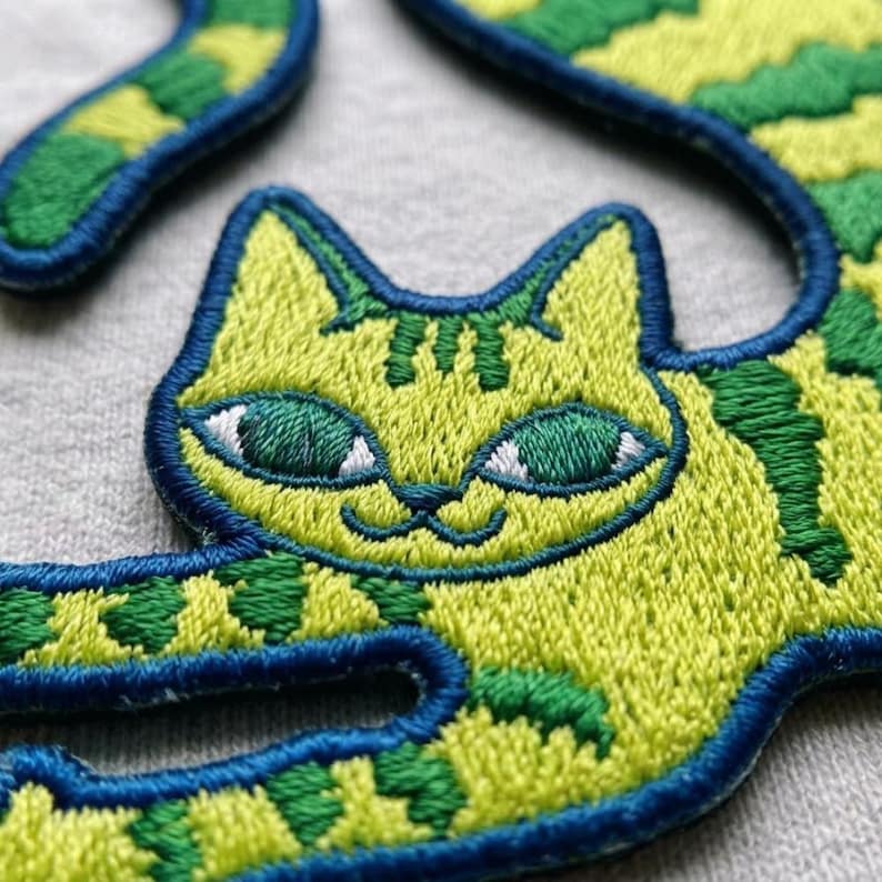 May include: An embroidered patch featuring a stylized cat with lime green fur and dark green stripes. The cat has large, almond-shaped green eyes with white accents and a dark blue stitched border that outlines the entire shape of the feline figure.