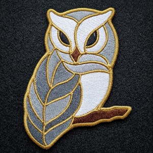 May include: An embroidered owl patch featuring a stylized design with white, grey, and brown thread. The owl has large dark eyes, a brown beak, and is perched on a brown branch. The entire design is outlined with a thick, metallic gold thread.