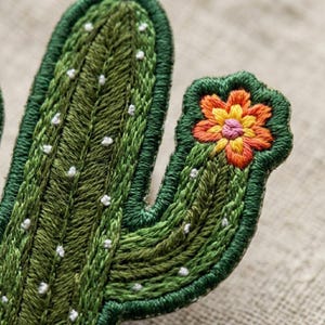May include: A cactus-shaped embroidered patch featuring dark and light green thread detailing with small white stitched dots representing spines. A vibrant orange and yellow flower with a pink center is stitched onto the cactus arm.