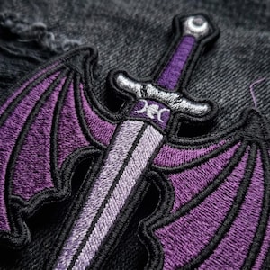 May include: An embroidered patch featuring a sword with large, purple bat-like wings extending from the sides. The sword handle is purple with a silver crossguard, and a triple moon symbol is stitched in silver thread at the center of the hilt.