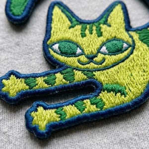 May include: An embroidered patch featuring a lime green cat with dark green stripes and large green eyes. The cat is depicted in a reclining position with its front paws extended forward. The entire design is outlined with a thick dark blue border.