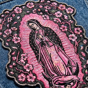 May include: An embroidered patch featuring a figure with hands clasped in prayer, surrounded by a vibrant pink aura and a dark border adorned with pink cherry blossoms. The figure wears a dark cloak over a pink robe, set against blue denim fabric.