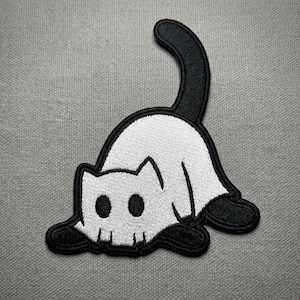 May include: A black and white embroidered patch featuring a stylized cat in a pouncing position. The cat has a white body with black circular eyes, black paws, and a long, curved black tail, all outlined with a thick black border.