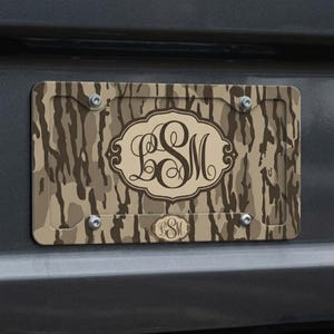 May include: License plate with a camouflage pattern in shades of brown and tan. The plate features a decorative oval with the initials "LSM" in a stylized font. A smaller version of the initials is at the bottom.