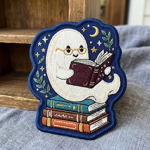 May include: An embroidered patch featuring a white ghost wearing gold glasses and reading a dark red book. The ghost sits atop a stack of three books in teal, brown, and orange. The dark blue background includes yellow stars, a crescent moon, and green leaves.