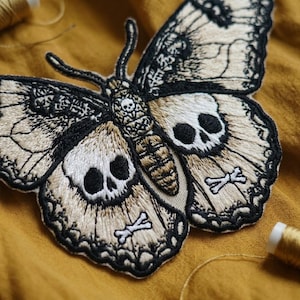 May include: An embroidered patch shaped like a moth with beige and black thread. The wings feature skull patterns and small crossbones, while the central body displays a skull design. The embroidery is detailed with black outlines and intricate textures.