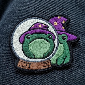 May include: An embroidered patch featuring a green frog wearing a purple wizard hat with yellow stars. The frog is positioned inside a white and gray crystal ball with a brown base. A second green frog in a purple hat is visible to the right side.