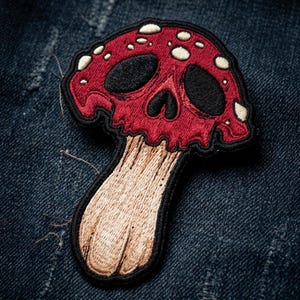 May include: An embroidered patch featuring a red mushroom cap shaped like a skull with black eye sockets and a nasal cavity. The cap has small white spots, and the mushroom stem is light brown with vertical stitching details and a black border.